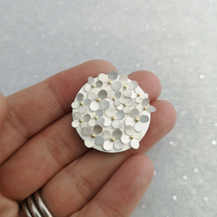 a hand holding a round silver brooch covered in flowers