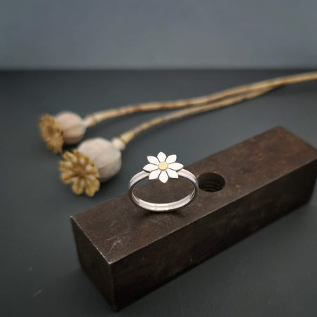 silver and 18ct gold band with a tiny dahlia on top, sitting on top of a block of rusty meatl, with seed pods in the background